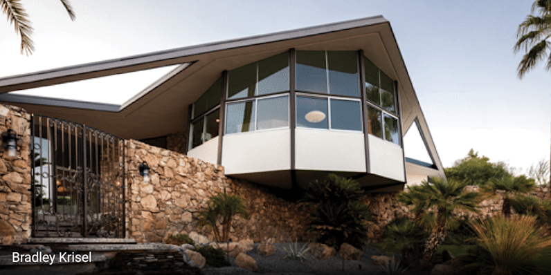Mid-century modern home in Palm Springs, California featuring dramatic angular rooflines, cantilevered design, large glass windows, and stone exterior walls, surrounded by palm trees and desert landscaping under bright sunlight.