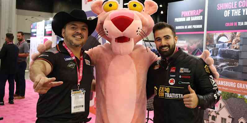 Two men pose with the Pink Panther mascot at a tradeshow. The man on the left wears a black cowboy hat and a black polo shirt with sponsor logos. He gestures with a peace sign. The man on the right wears a black jacket with sponsor logos and gives a thumbs up. The Pink Panther stands between them with its arms around their shoulders. Partially visible behind them are trade show booths with signage about roofing and insulation products. A pink carpet runner is visible below.