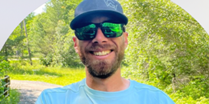 Person wearing a blue cap and light blue shirt standing outdoors on a sunny day with trees and a path in the background.