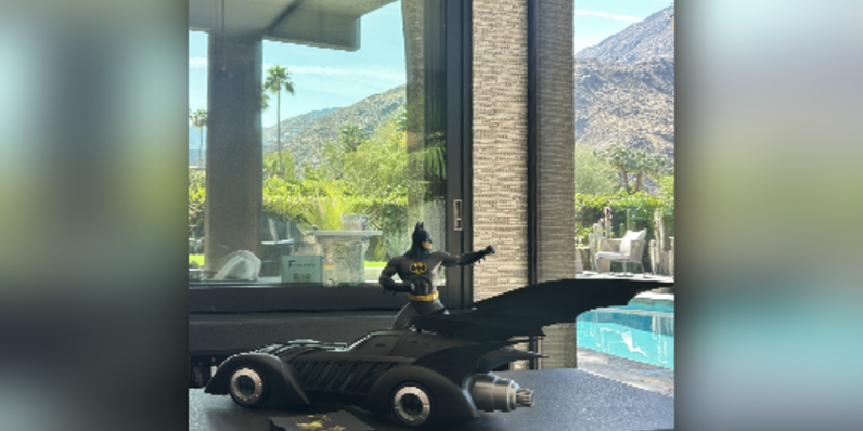 Modern living room with floor-to-ceiling glass windows offering panoramic views of desert mountains and cactus landscape, featuring gray sectional sofa with patterned and yellow accent pillows, sleek black fireplace, and minimalist decor.