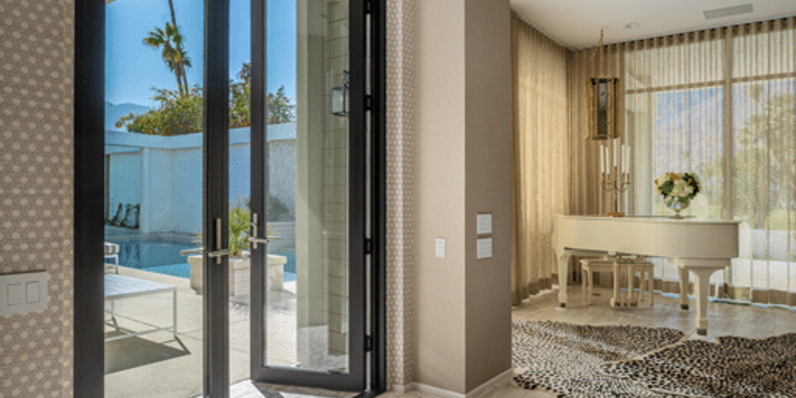 Modern interior entryway with large black-framed glass pivot doors opening to a sunlit patio with swimming pool, featuring light wood flooring, neutral walls, and a white grand piano accented by a patterned area rug.