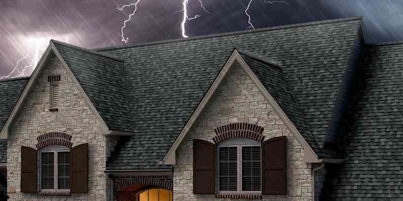 Photo of the top of a roof with gray shingles set against a dark, stormy sky with lightning