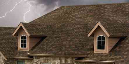 Tan colored house with brown shingles set against a dark, stormy sky with lightning flashing