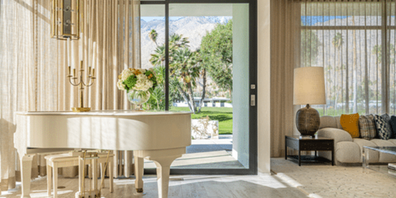 Elegant living room with floor-to-ceiling glass doors opening to a patio with mountain views, featuring a white grand piano, modern chandelier, neutral-toned sofa, patterned area rug, and sheer curtains allowing natural light.