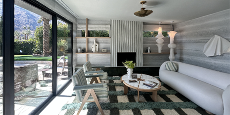 Modern living room with floor-to-ceiling sliding glass doors opening to a patio with outdoor seating, featuring a white curved sofa, two light wood armchairs, round coffee table, and striped green and beige area rug under a wood-paneled ceiling.