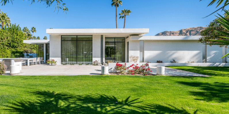 Mid-century modern home with flat roof design, white stucco exterior, and expansive floor-to-ceiling glass doors opening to a large patio, surrounded by manicured green lawn, palm trees, and mountain views under a clear blue sky.