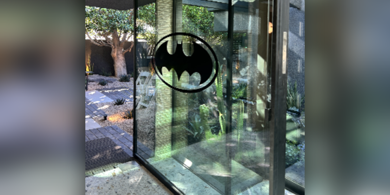 Glass pivot door with black metal frame featuring a Batman logo decal, opening to a modern indoor-outdoor space with concrete flooring, landscaped garden, and natural light reflections.