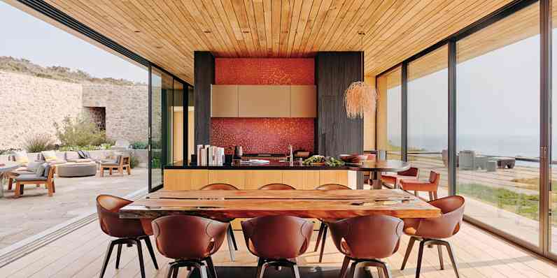 Minimalistic living area with table and chairs and two walls of floor to ceiling windows that are open, leading to a patio.