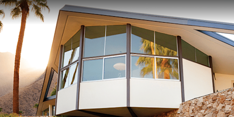 Mid-century modern home in Palm Springs, California featuring dramatic angular rooflines, cantilevered design, large glass windows, and stone exterior walls surrounded by palm trees and desert landscaping.