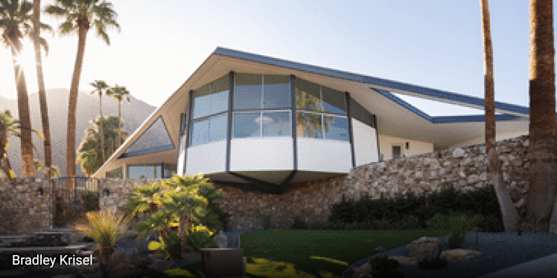 Mid-century modern home in Palm Springs, California featuring dramatic angular rooflines, cantilevered design, large glass windows, and stone exterior walls, complemented by desert landscaping with palm trees and gravel pathways.