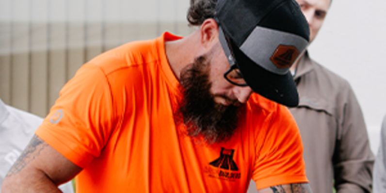 Person in an orange work shirt and black cap leaning forward while working on a project, with others observing in the background.