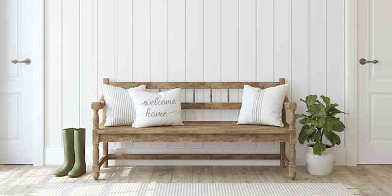 Rustic wood bench with white throw pillows shown in front of a white slatted wall, green plant in a white pot and tall green rainboots next to the bench.