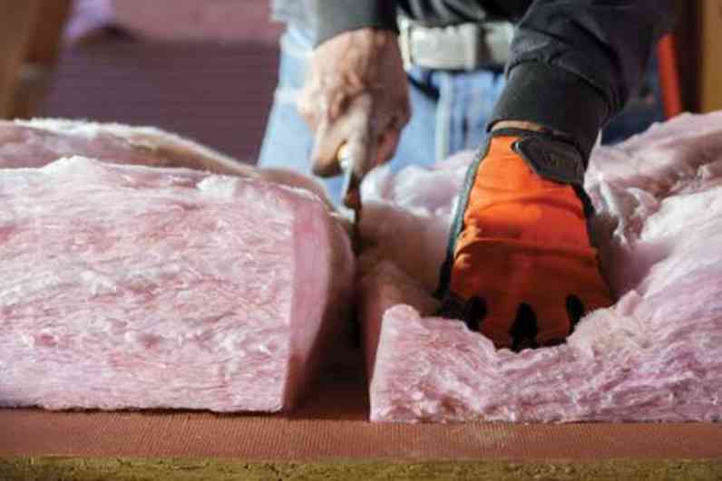 A professional insulation installer cuts PINK Next Gen™ Fiberglas™ with a large shear while wearing orange gloves.
