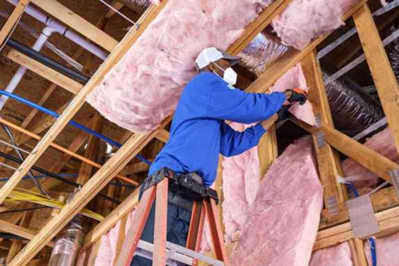A professional installer stands on a ladder, reaching up into the ceiling to install and lay down PINK Next Gen™ Fiberglas™ batts into the ceiling cavities.