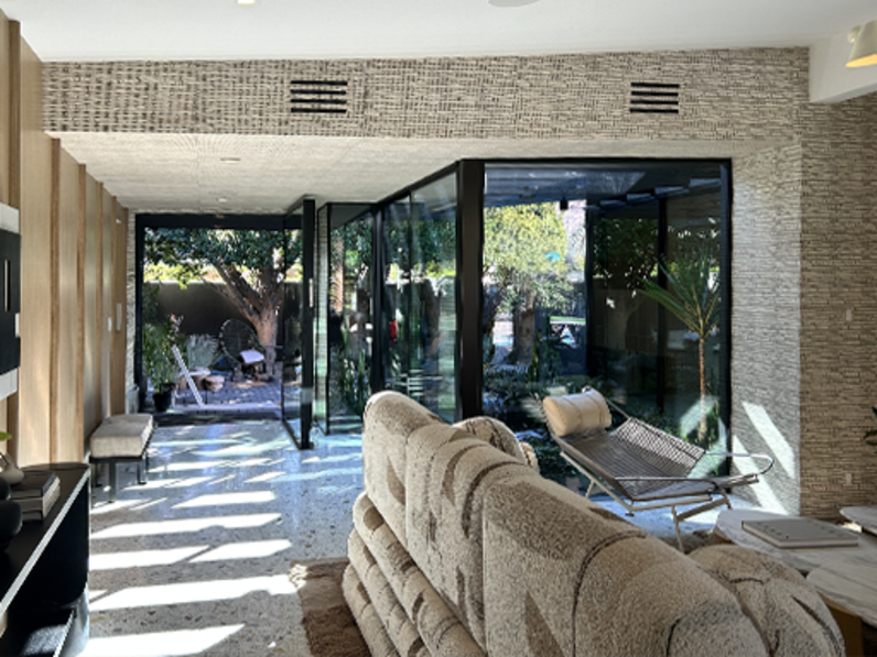 Modern living space with textured stone walls, large glass pivot doors opening to a landscaped courtyard, plush beige sectional sofa, minimalist lounge chair, and abundant natural light creating an indoor-outdoor connection.