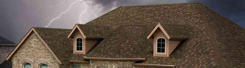 Tan colored house with brown shingles set against a dark, stormy sky with lightning flashing