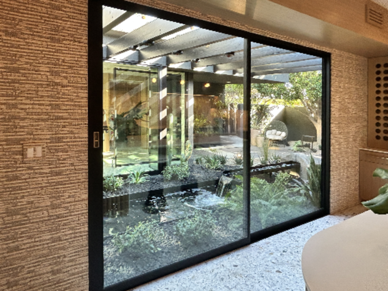 Modern interior with large sliding glass doors offering a view of a landscaped courtyard featuring a water feature, greenery, and a pergola structure, complemented by textured wall finishes and natural light.