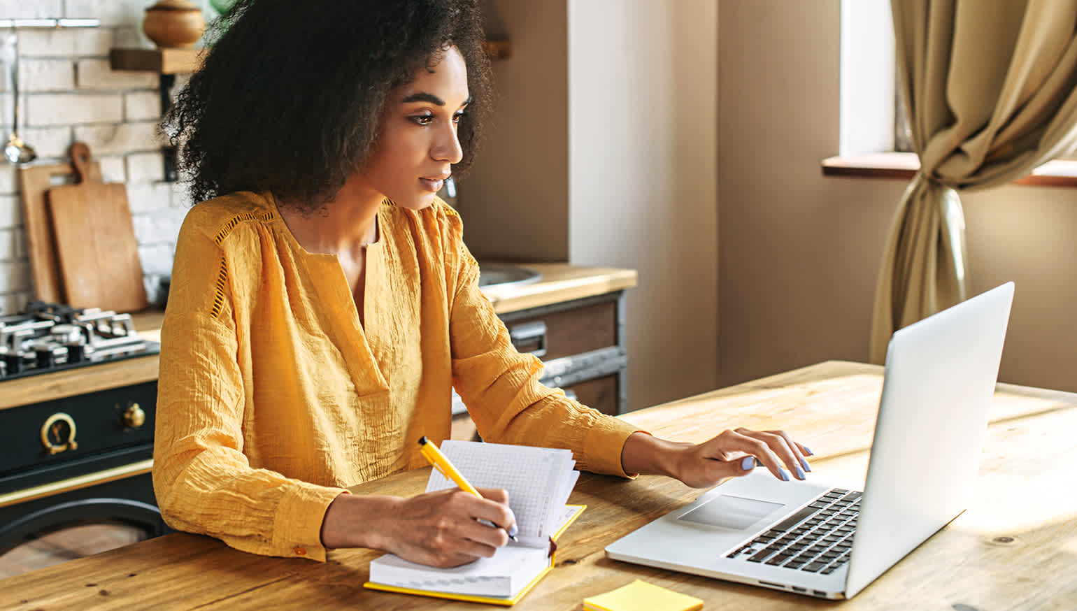 Photo of a woman sitting at a kitchen table with a laptop open, taking notes in a notebook