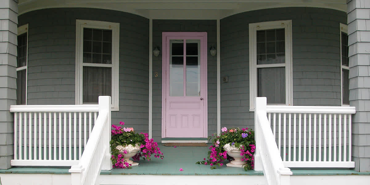 Gray house with pops of color: pink door, green porch, bright pink flowers, and white railing around the porch and stairs
