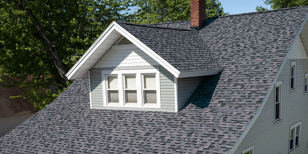 Light blue-grey house with a dormer with three windows and white scalloped siding and a purple-hued asphalt shingle roof from Owens Corning, Midnight Plum.