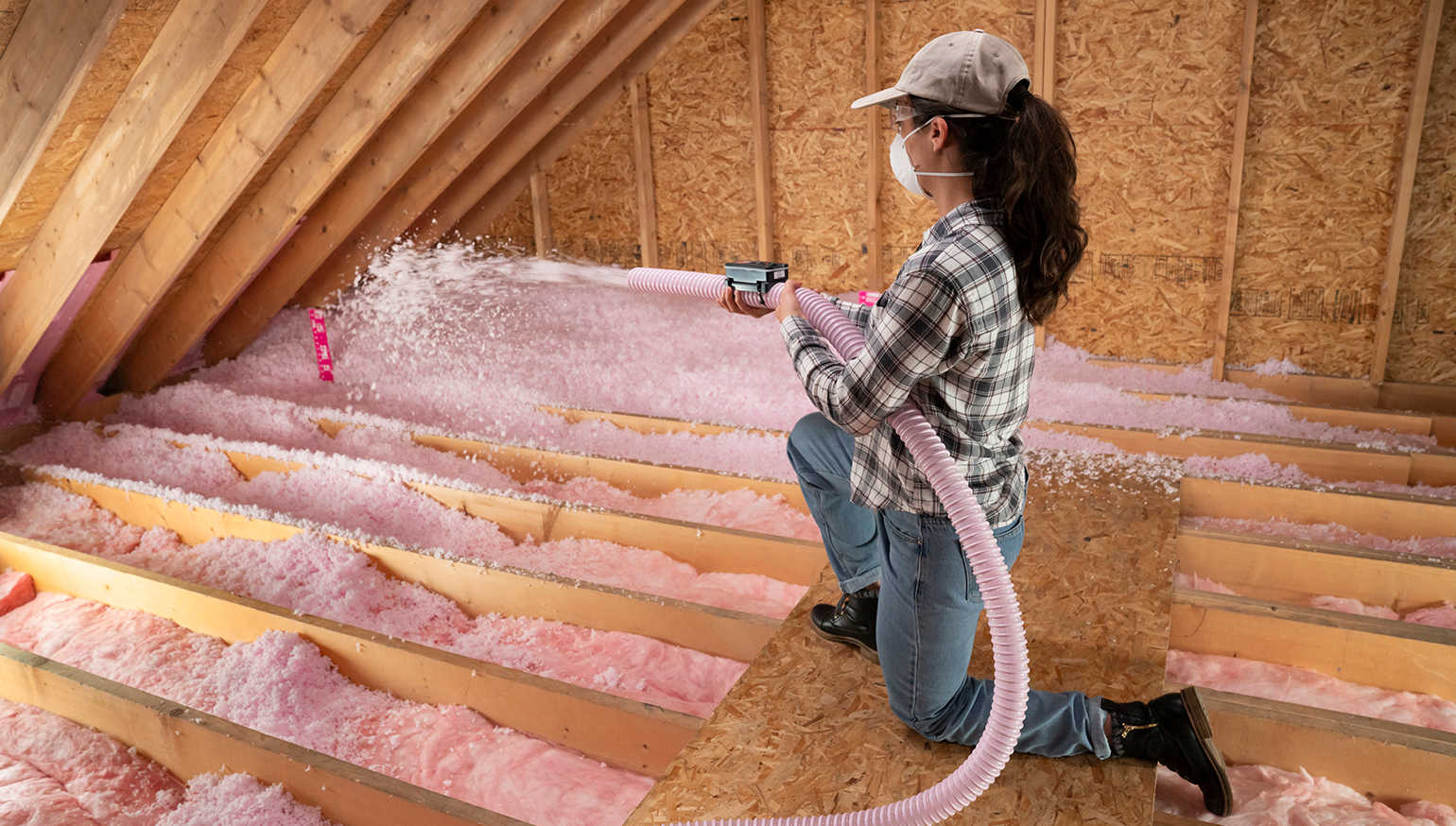 Women in plaid shirt using Owens Corning Atticat machine to blow in pink insultation to attic flooring