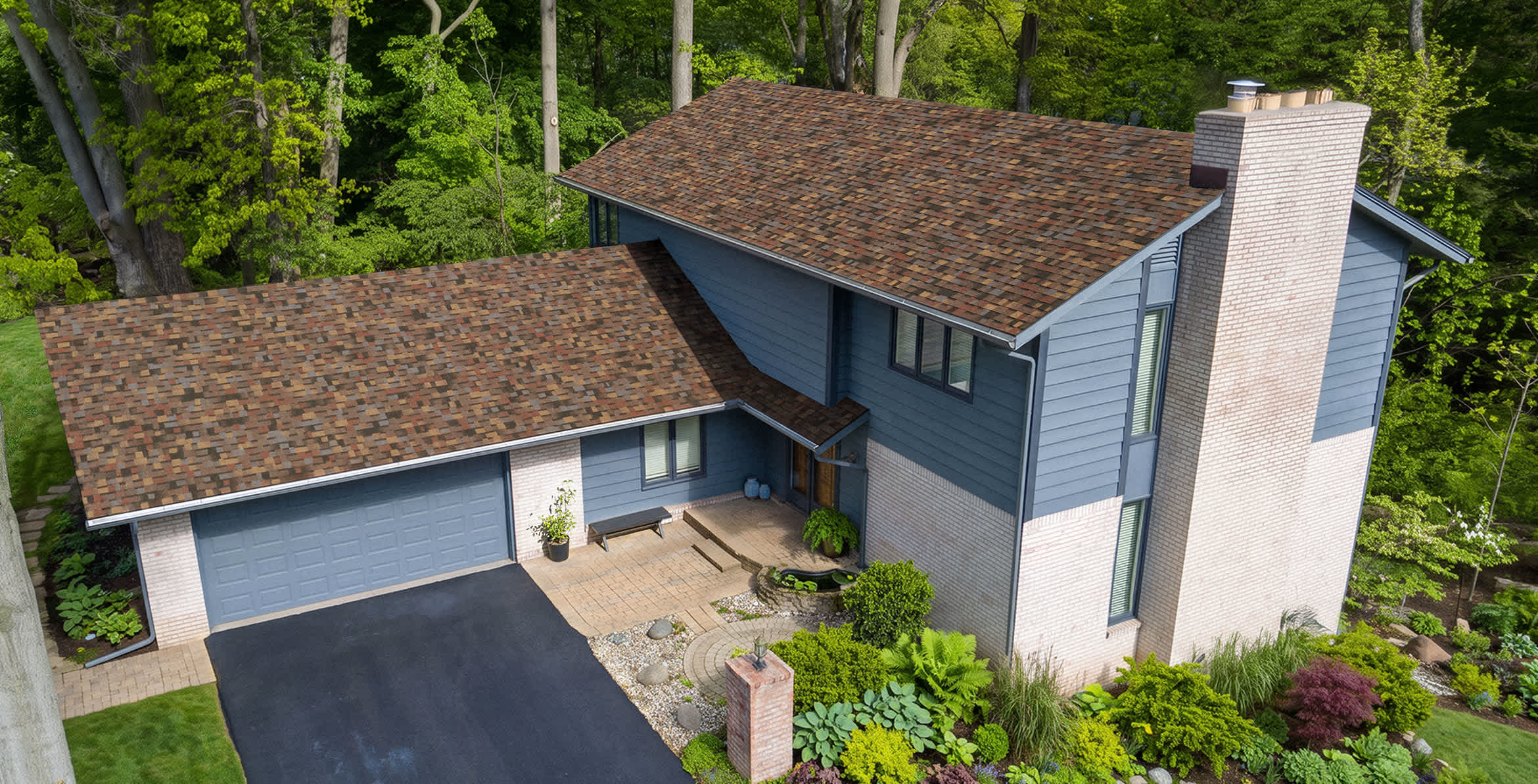 A blue sided house with tan brick accents sits on a beautifully landscaped yard with new Summer Harvest roof