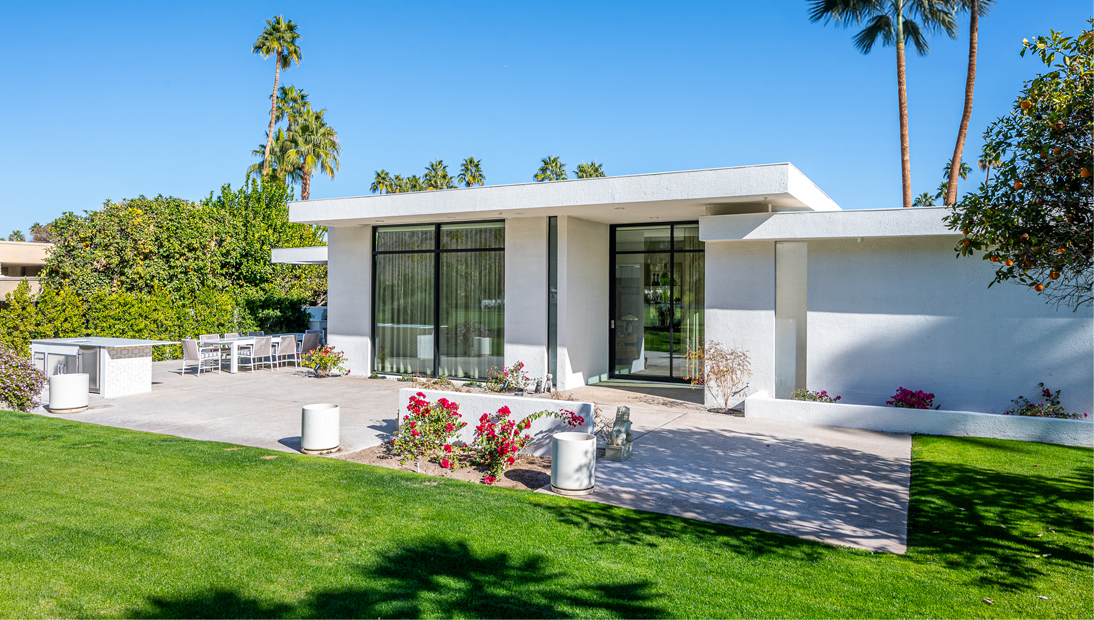 Mid-century modern home with flat roof design, white stucco exterior, and large floor-to-ceiling glass windows, featuring an outdoor patio with dining area and landscaped lawn surrounded by palm trees and flowering plants.