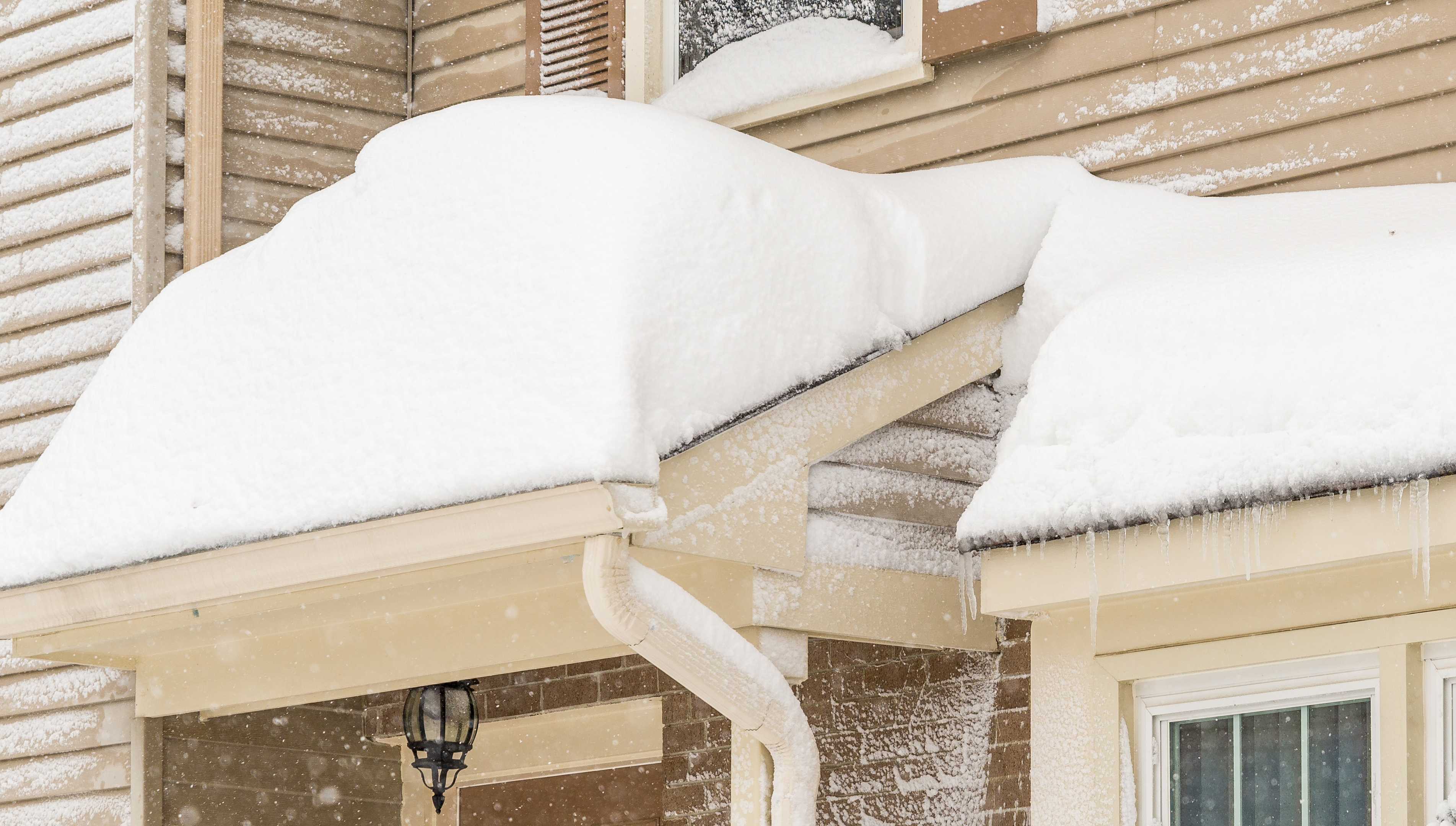 Tan sided home with several feet of snow built up on the porch overhang and window dormers.