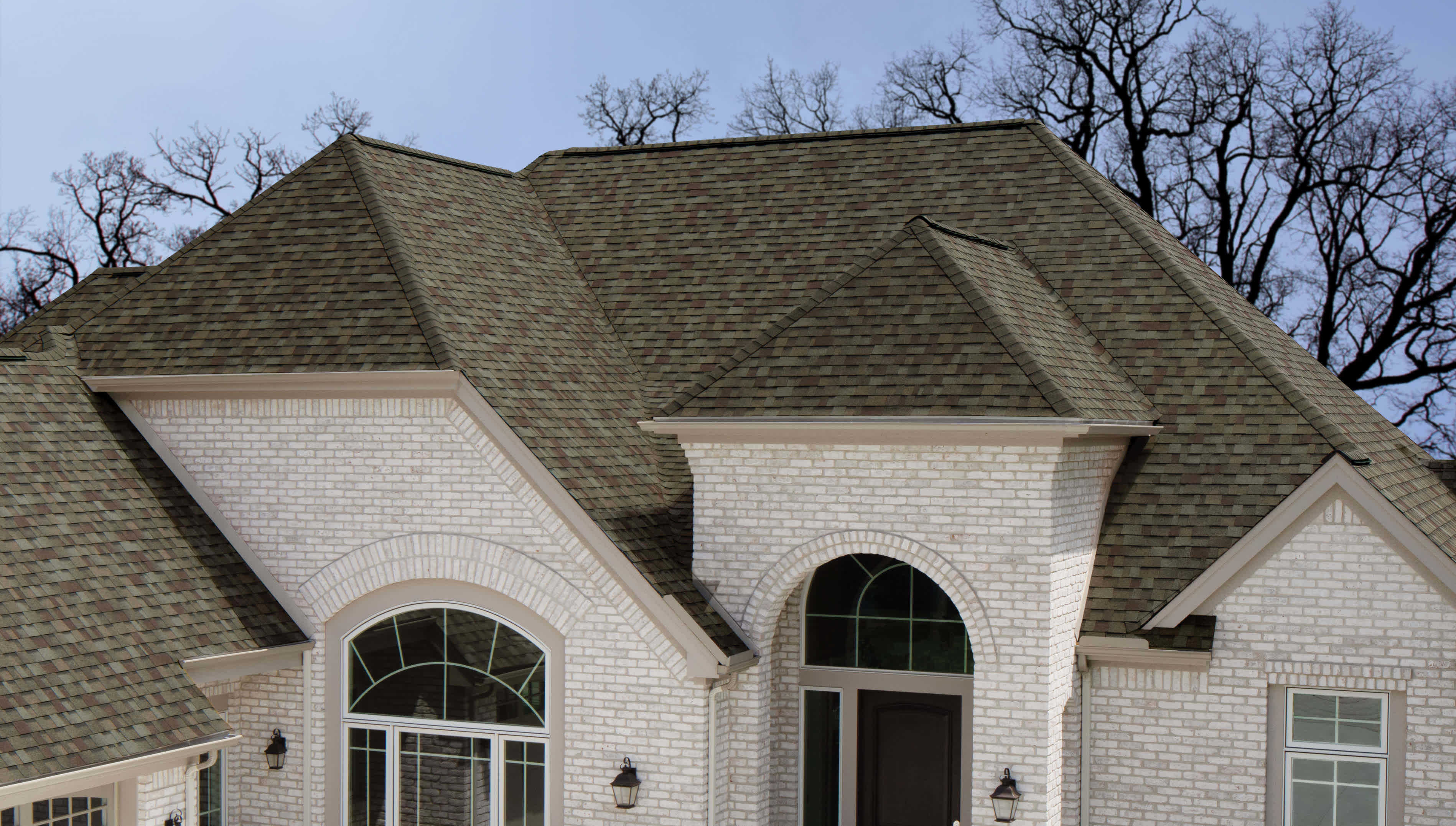 Tan brick home with a brand new roof, and trees in background without any leaves.