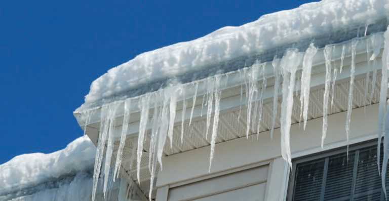 A corner angle of roof and gutters built up with snow and ice damming with long icicles hanging from the gutters.