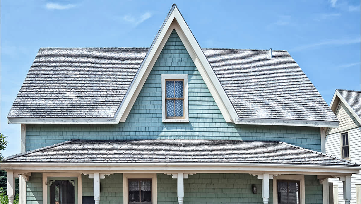 Front view of a blue shake house with high peak dormer, and old worn grey shingle roof