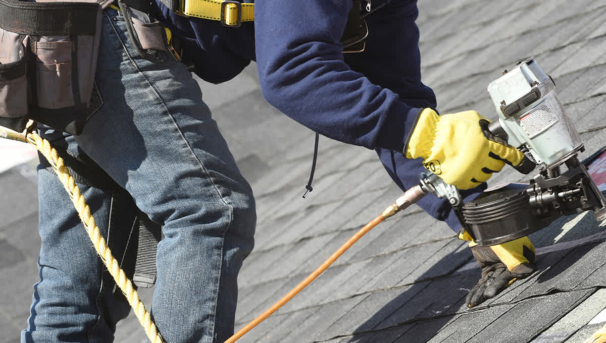 Close up view of a roofer using a nail gun to install grey shingles on a roof