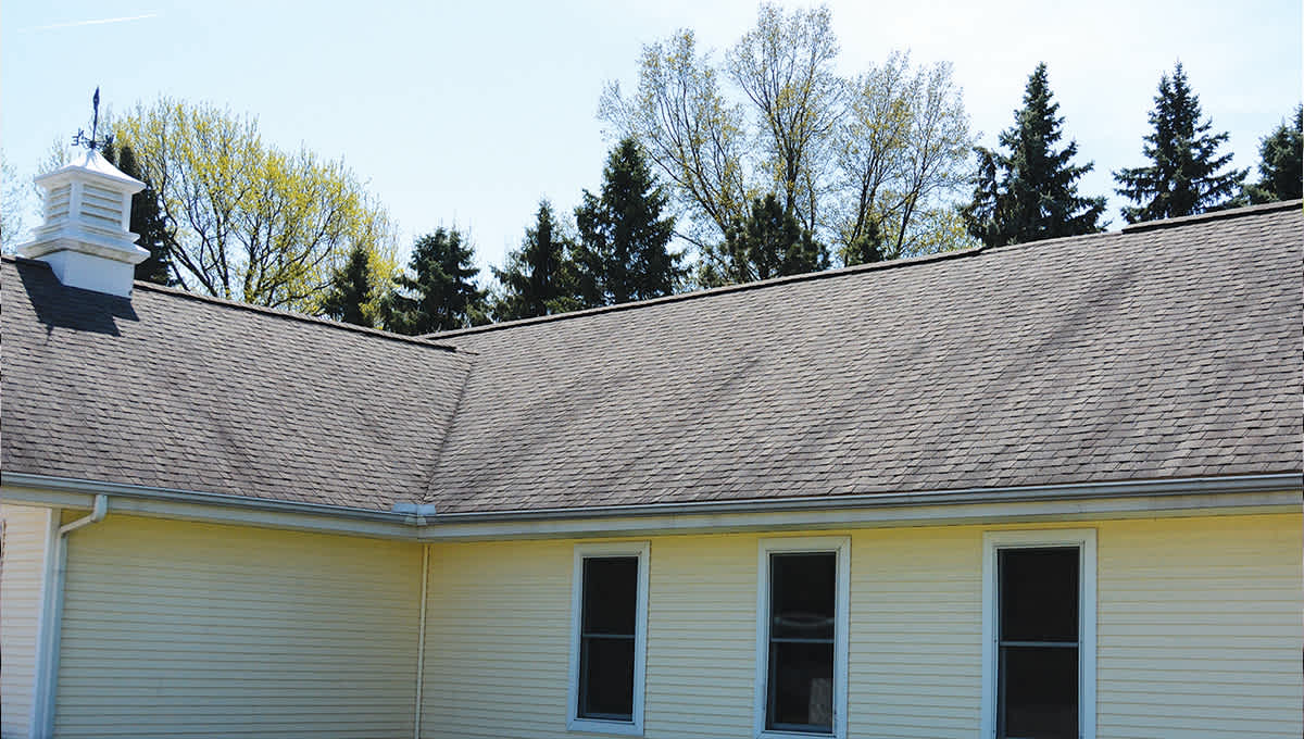 House with pale yellow siding, three white windows and a brown asphalt shingle roof with dark streaks