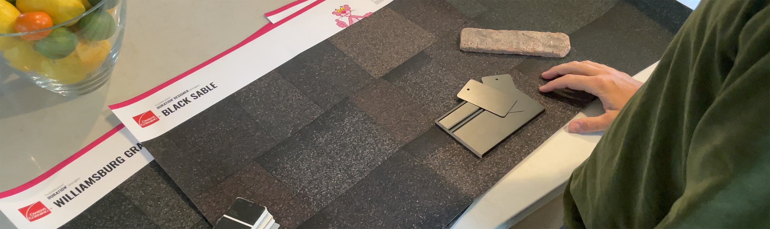 Three Owens Corning printed shingle posters spread across the kitchen counter with pantone swatches, paint chips, and brick veneers