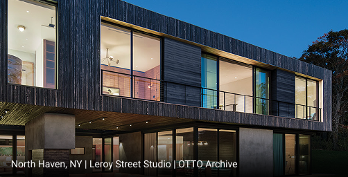 Contemporary two-story home in featuring dark vertical wood siding, cantilevered upper level with floor-to-ceiling glass windows, and open-concept lower level with sliding glass doors overlooking landscaped lawn.