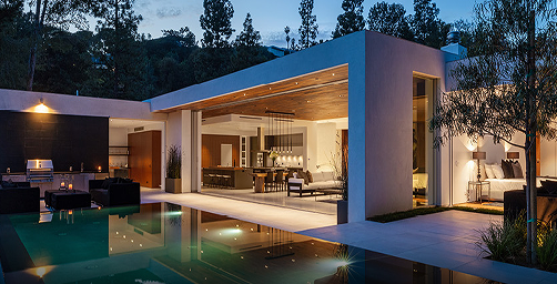 Luxury modern home with open-concept design featuring floor-to-ceiling sliding glass doors, white stucco exterior, wood-paneled ceiling, and outdoor living space with illuminated swimming pool and lounge seating at dusk.