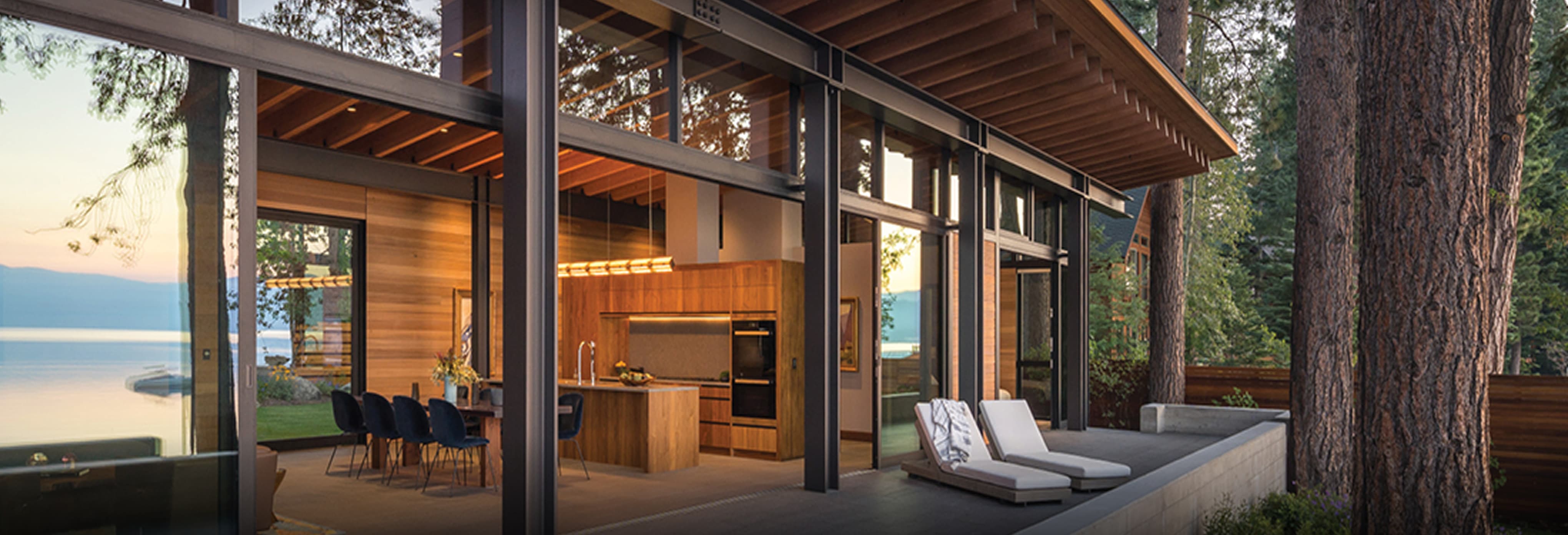 Modern lakefront home with exposed wood ceiling beams, floor-to-ceiling glass walls, open-concept kitchen with wood cabinetry, and outdoor patio featuring lounge chairs overlooking serene water views.