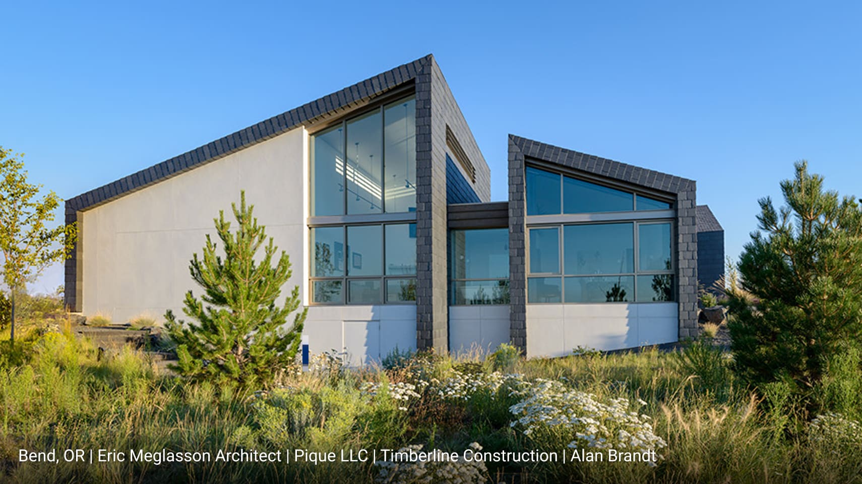 Modern home in Bend, Oregon featuring angled rooflines, gray stone tile cladding, large floor-to-ceiling glass windows, and minimalist landscaping with native plants under a clear blue sky.