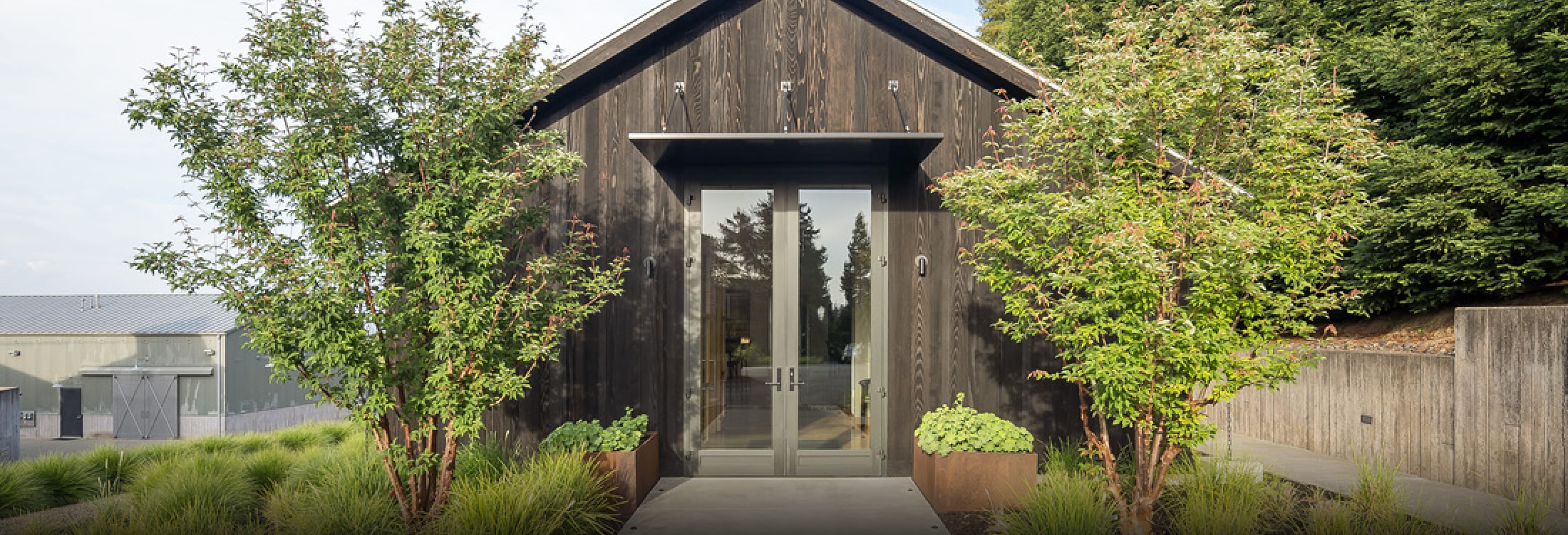 Contemporary home exterior with dark wood siding, pitched roof, and glass front door framed by minimalist metal awning, surrounded by lush green trees and landscaped entryway.