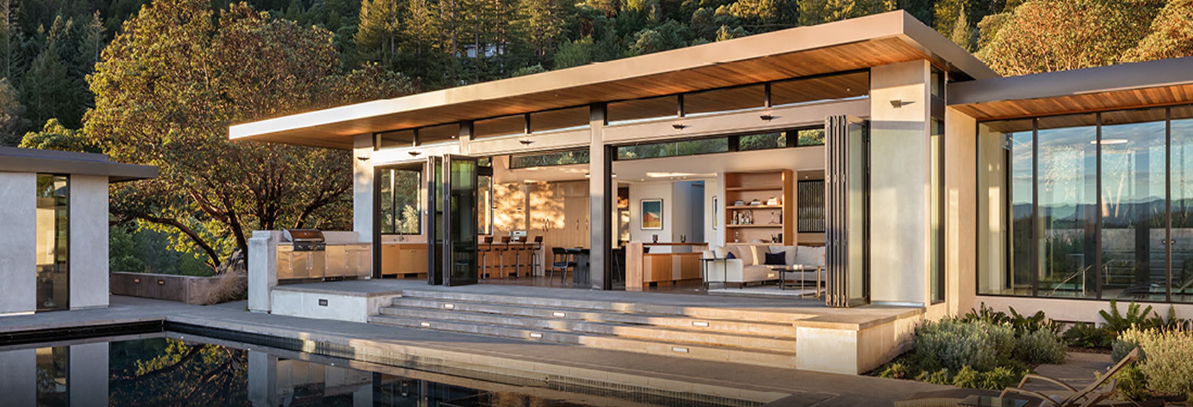 Luxury modern home with open-concept design featuring folding glass doors, natural wood ceiling, and outdoor living space overlooking a pool, surrounded by lush greenery and hillside views.