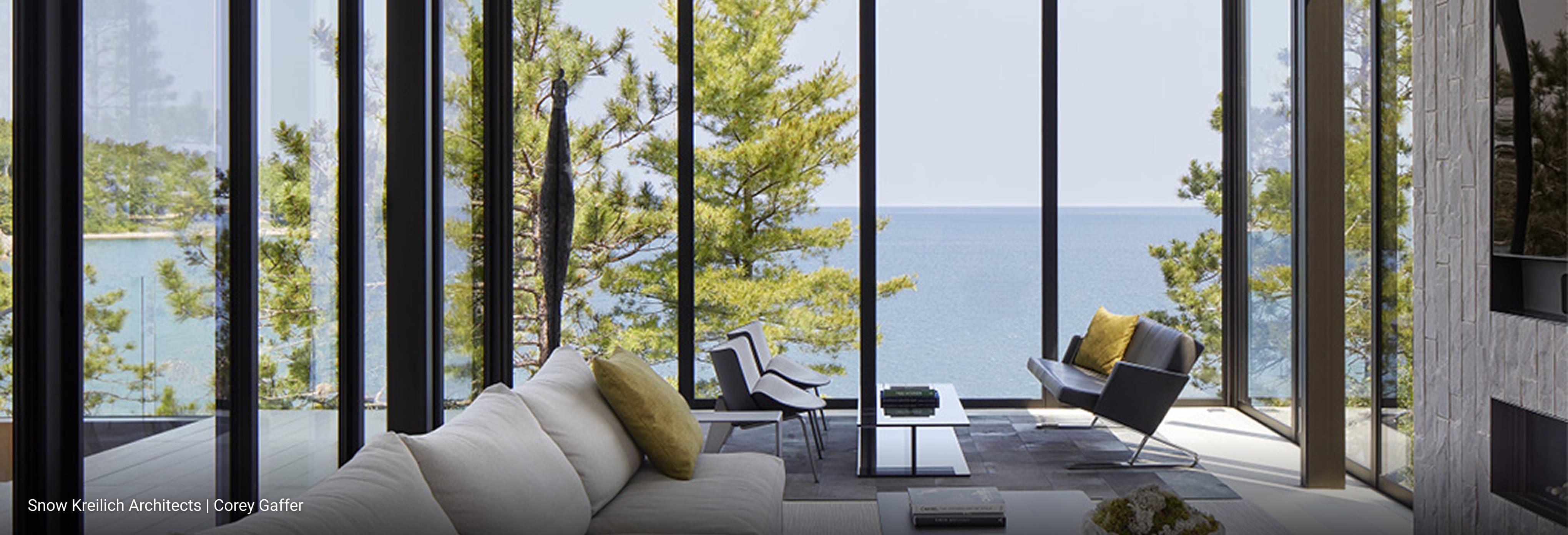 Modern living room with floor-to-ceiling glass walls offering panoramic lake views, minimalist furniture including light beige sectional sofa and black lounge chair, sleek coffee tables, and natural light highlighting contemporary design.
