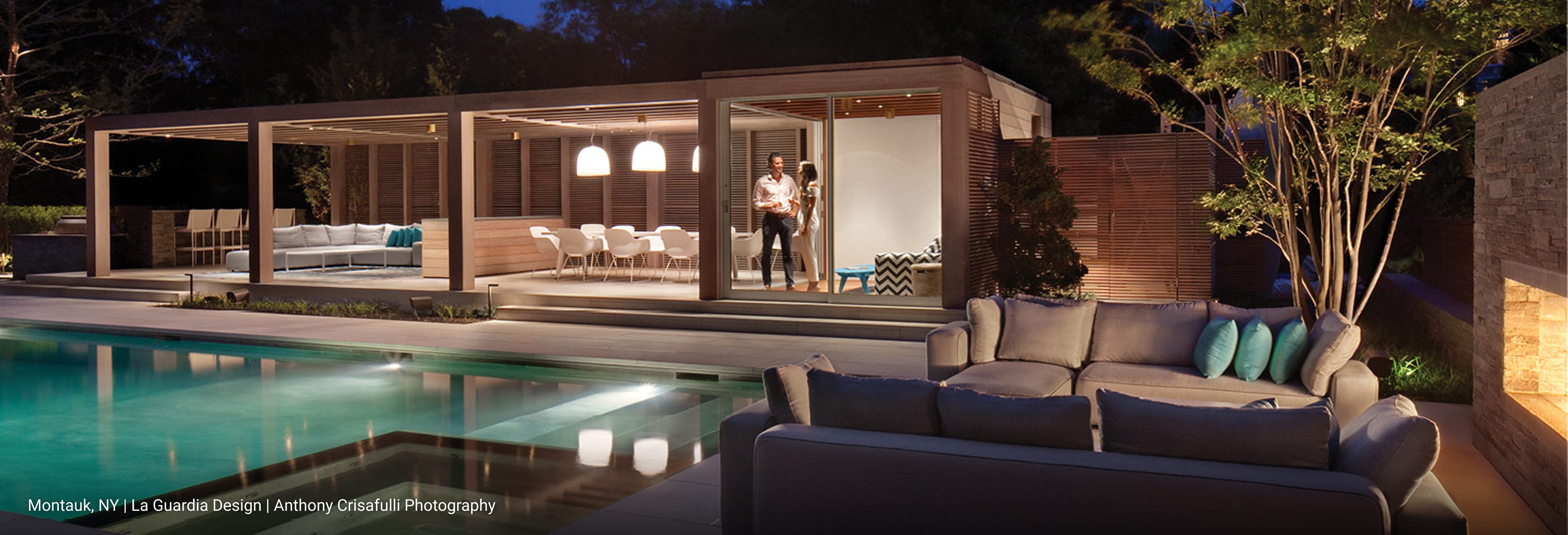 Luxury outdoor living space in Montauk, New York featuring modern pergola with integrated seating, dining area with pendant lights, and open-concept design overlooking a rectangular swimming pool, complemented by contemporary lounge furniture and accent pillows.