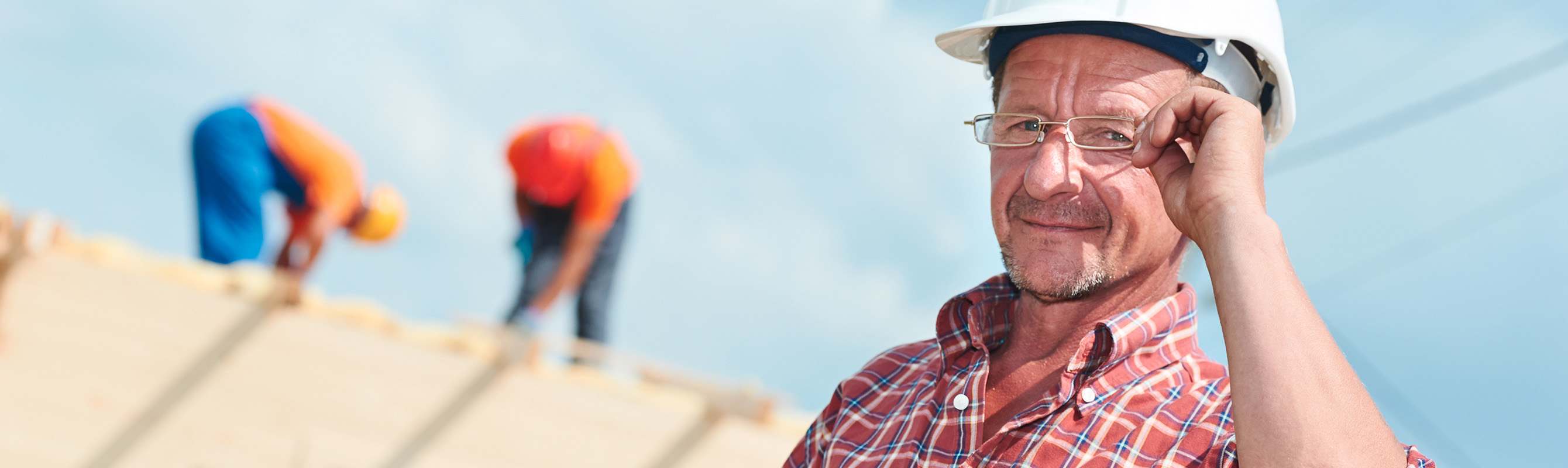 Close up of man wearing a white hard hat, adjusting glasses on his face, with blurred roofers on top of roof in background.