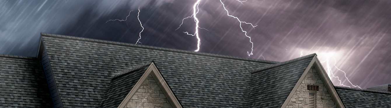 Photo of the top of a roof with gray shingles set against a dark, stormy sky with lightning