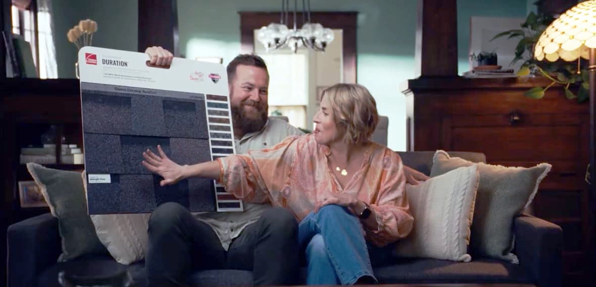 Erin and Ben Napier sitting on a couch looking at an Owens Corning Midnight Plum shingle sample board.
