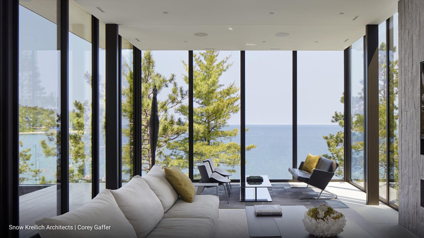 Modern living room with floor-to-ceiling glass walls offering panoramic lake views, minimalist furniture including light beige sofa and black lounge chair, sleek coffee tables, and natural stone accent wall.