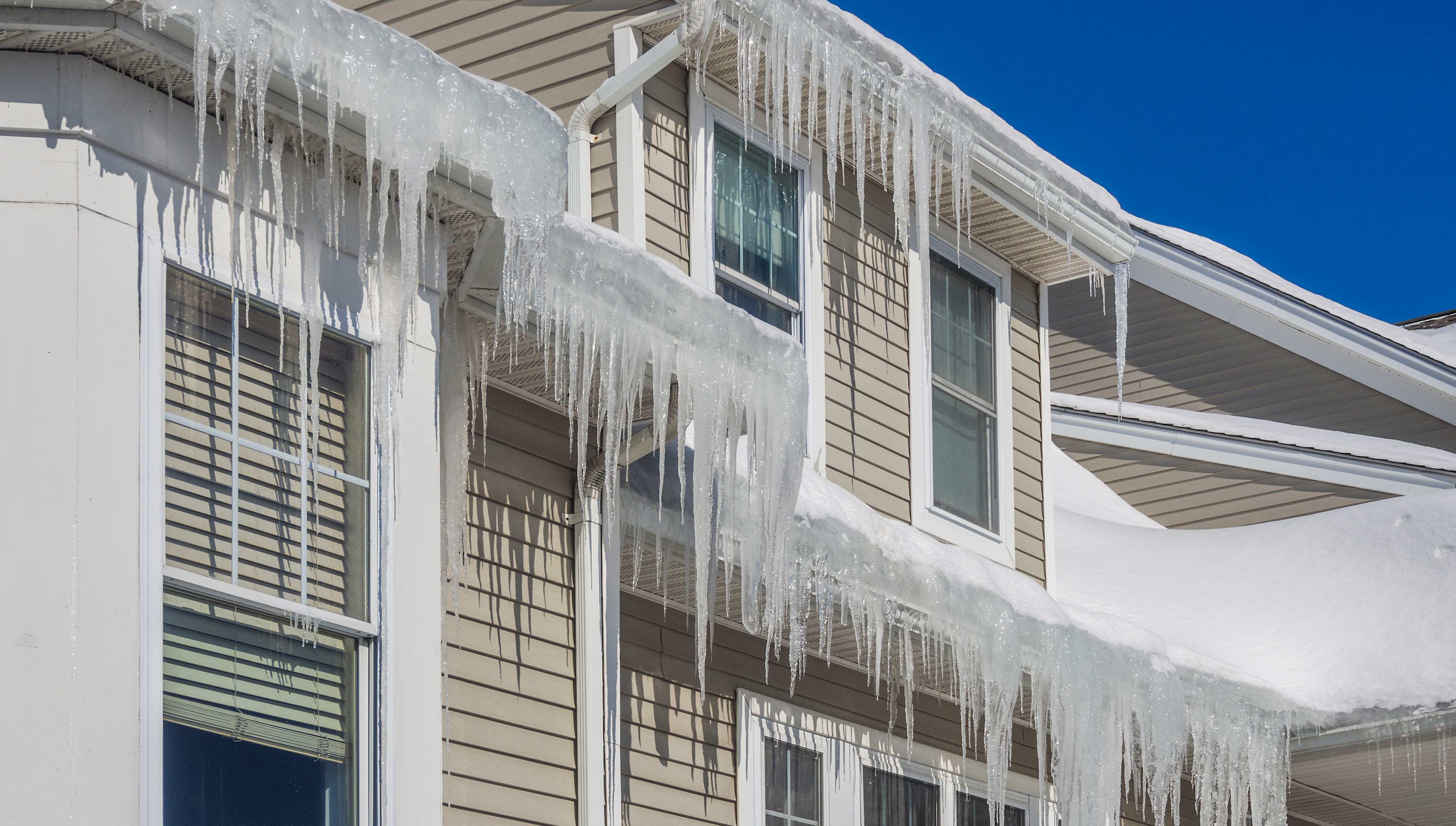 Tan house with snow covered roof and severe ice dams overhanging the gutters
