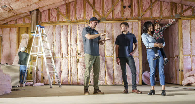 A mom and a dad stand with their daughter inside their home that is in the process of being built, while talking to their professional contractor and the little girl points at the PINK Next Gen™ Fiberglas™ that has been placed in the wall cavities.