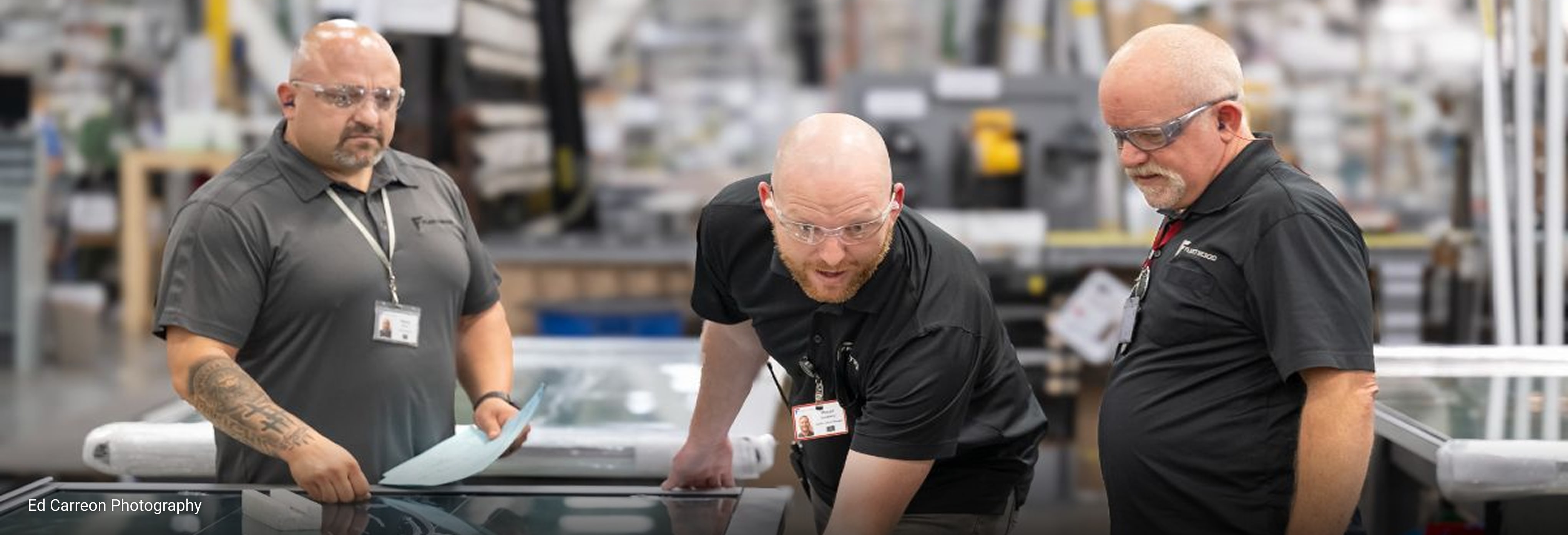 Three men in a warehouse, wearing Fleetwood-branded shirts, name badges, and safety goggles, working on a large pane of glass.