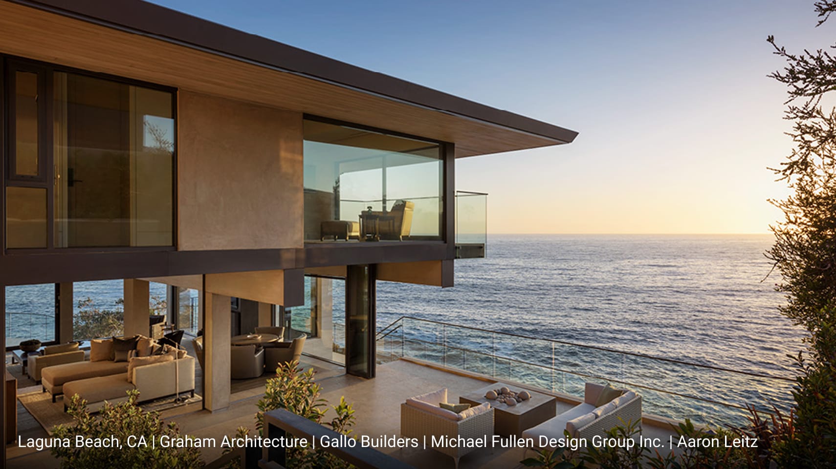 Luxury modern home in Laguna Beach, California featuring cantilevered upper level with glass balcony, expansive floor-to-ceiling windows, and open-concept outdoor living space overlooking the Pacific Ocean at sunset.